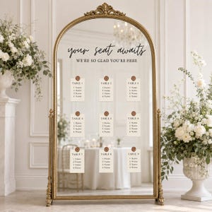 May include: An arched floor mirror with a gold frame featuring the black text "your seat awaits" and "WE'RE SO GLAD YOU'RE HERE". Nine cream-colored cards are attached to the glass, each labeled with a table number and four name placeholders.
