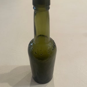 Könnte beinhalten: Eine antike olivgrüne Glasflasche mit dunklem Verschluss. Die Flasche hat einen abgerundeten Boden und einen langen Hals. Geprägter Text auf der Flasche lautet "J.C. B". Die Flasche stammt wahrscheinlich aus dem frühen 20. Jahrhundert.