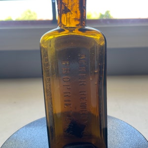 Peut inclure: Une bouteille en verre brun foncé avec un corps carré et un col étroit. La bouteille porte une inscription en relief qui dit "ACKER'S REMEDY FOR  PRURIE TOX"