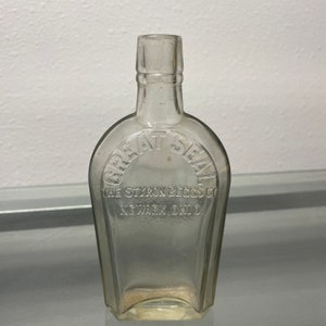 Pode incluir: Uma garrafa de vidro transparente com corpo retangular e topo arredondado. As palavras "GREAT SEAL" estão arqueadas no topo, com "THE STYRON BECGS CO" e "NEWARK, OHIO" abaixo. A garrafa está vazia.