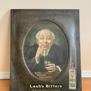 May include: Vintage metal sign advertising Lash's Bitters, a natural tonic laxative. The sign features a man in a suit holding a glass of the bitters and the text "Acts Without Discomfort".