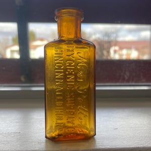 Könnte beinhalten: Eine kleine, bernsteinfarbene Glasflasche mit quadratischer Form und abgerundetem Oberteil. Die Flasche hat einen geprägten Text mit der Aufschrift "M.J.  Peters Hygienic Supply Co. Cincinnati, Ohio, Inc."