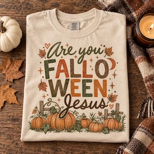May include: A beige crew neck t-shirt featuring a graphic print with the text Are you FALLO WEEN Jesus. The design includes orange and green autumn leaves, small stars, hearts, a crescent moon, and a pumpkin patch with a wooden fence.