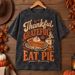 Puede incluir: Una camiseta gris carbón con efecto lavado al ácido que presenta un estampado gráfico con el texto Thankful GRATEFUL & EAT PIE. El diseño incluye una porción de tarta de calabaza en un plato, una taza de café y hojas otoñales en tonos naranja, rojo y marrón.