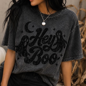 May include: A charcoal grey acid-wash t-shirt featuring a black graphic print with the text "Hey Boo" in a cursive font. The design includes two ghost illustrations, a crescent moon, and several small four-pointed stars surrounding the text.