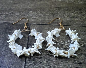 May include: A pair of white hoop earrings. Each earring is made of small, bone-shaped pieces arranged in a circular pattern. The earrings have gold-colored ear wires.