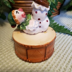 May include: A ceramic clown figurine with a brown drum base. The clown is wearing a green hat, white ruffled collar, and white outfit with red polka dots. The drum is tan with brown trim.