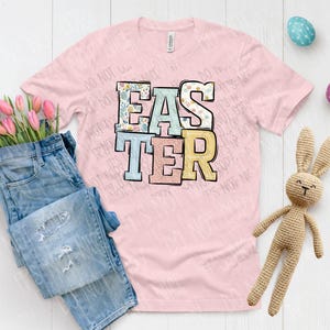 以下が含まれることがあります： パステルカラーの「イースター」のグラフィックがプリントされたピンクのTシャツ。グラフィックは、さまざまな模様の文字で構成されています。Tシャツは、白い木製の表面に、青いジーンズ、かぎ針編みのウサギ、イースターエッグと一緒に置かれています。