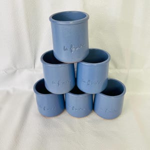 May include: A stack of six blue ceramic pots arranged in a pyramid formation. Each cylindrical pot features a smooth, matte finish and the embossed text la fermière on the front. The pots have a simple, open-top design with a slightly flared rim.