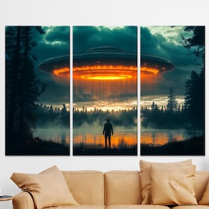 May include: A triptych canvas print depicts a UFO hovering over a lake with a person standing on the shore. The scene is set at dusk, with an orange glow emanating from the spacecraft and reflecting on the water. Dark trees frame the scene.