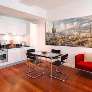 Puede incluir: Un comedor moderno con una cocina blanca y una gran impresi&oacute;n de paisaje des&eacute;rtico colgada en la pared. La mesa de comedor es de vidrio y tiene cuatro sillas negras. Un sill&oacute;n rojo con un coj&iacute;n beige est&aacute; en la esquina de la habitaci&oacute;n.