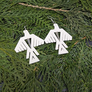 May include: A pair of white, geometric bird-shaped earrings with silver hooks. The earrings are made of wood and have a cutout design.