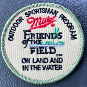 May include: Circular patch with the text "Outdoor Sportsman Program" and "Miller" in red script. The patch also reads "Friends of the Field" and "On Land and in the Water". The patch has a green border.