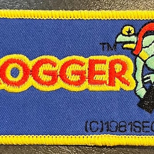May include: Embroidered patch featuring the word "FROGGER" in red and yellow, with a green frog character carrying a briefcase. The patch has a blue background with a yellow border. Text at the bottom reads "(C)1981 SEGA".
