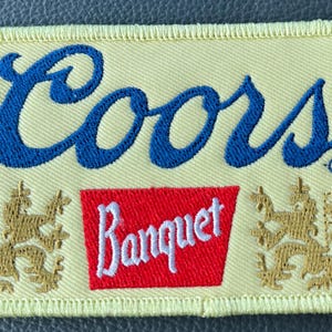 May include: Embroidered patch with the Coors logo in blue script above a red rectangle with "Banquet" in white script. The patch has a yellow background with gold lion emblems on each side.