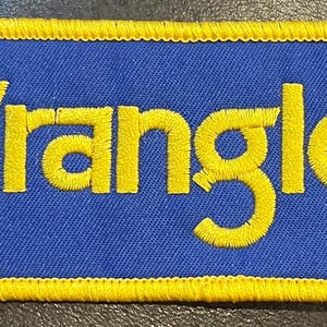 May include: Embroidered patch with the word "Wrangler" in yellow, stitched onto a blue rectangular background. The patch has a yellow border. This patch is likely for clothing or accessories.