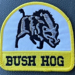 May include: Embroidered patch featuring a black and white illustration of a wild boar. The patch has a white background with a yellow border and the words "BUSH HOG" in black block letters.
