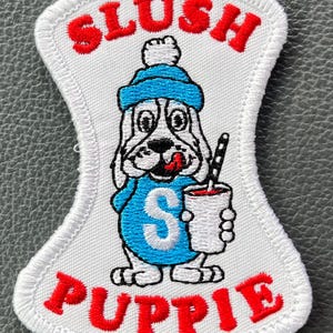 May include: White patch with a cartoon dog wearing a blue hat and sweater, holding a drink with a straw. The patch has the words "SLUSH" and "PUPPIE" in red, with a white border. The dog has a red tongue sticking out.
