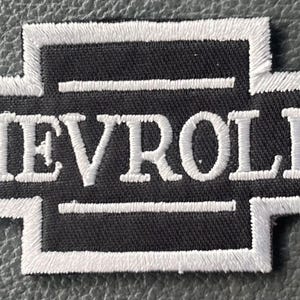 May include: Embroidered patch featuring the Chevrolet logo in a classic bowtie design. The patch has a black background with white embroidered lettering and border. The patch is rectangular and appears to be made of fabric.