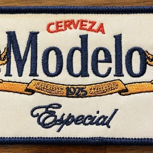 May include: A white embroidered patch with a blue border. The patch features the text "Cerveza Modelo Especial" in blue with a gold ribbon and two gold lions.