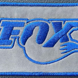 May include: Embroidered patch featuring the word "FOX" in blue, with a stylized design. The patch has a light gray background and a blue stitched border. Ideal for jackets, bags, or other apparel.