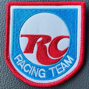 May include: Embroidered patch with a shield shape. The patch features a white circle in the center with red "RC" lettering. The background is blue with the words "RACING TEAM" in white. The border is red and white.