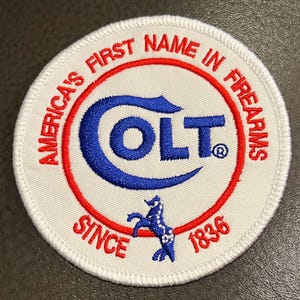 May include: A circular patch with the text "America's First Name in Firearms" in red, encircling a blue "COLT" logo. The patch includes a blue horse graphic and the text "SINCE 1836". The background is white.