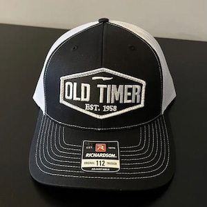 May include: Black and white trucker hat with a white embroidered patch that says "Old Timer Est. 1958". The hat has a white mesh back and a black front with a white adjustable strap.