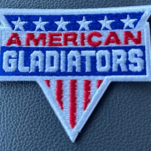 May include: Embroidered patch featuring the text "AMERICAN GLADIATORS" in red and blue, with white stars and stripes. The patch has a white border and a triangular bottom design. Ideal for jackets, bags, or other apparel.