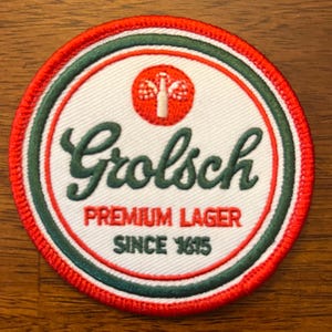 May include: A white, round, embroidered patch with a green and red border. The patch features the text "Grolsch Premium Lager Since 1615" and a red circle with a white bottle and green leaves.