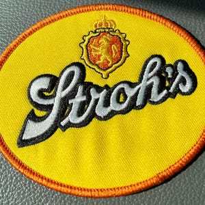 May include: Embroidered patch with the word "Stroh's" in a cursive font, outlined in black and white, on a yellow oval. The patch features a crest with a lion and crown, and an orange border.