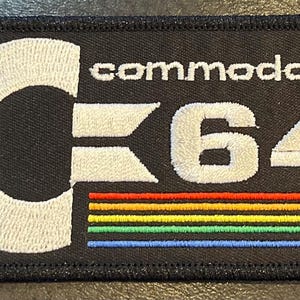 May include: Black embroidered patch featuring the Commodore C64 logo. The logo includes the word "commodore" above the "C64" text. The "C64" is white with a rainbow-colored stripe at the bottom. The patch has a black border.