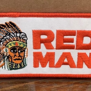 May include: A white embroidered patch with an orange border. The patch features a Native American man wearing a headdress and the text "RED MAN" in orange lettering with a registered trademark symbol.