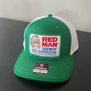 May include: Green and white trucker hat with a curved brim. The front features a patch with the text "RED MAN" and an illustration of a Native American chief. The hat also has the text "ALL AMERICAN PULLING SERIES".