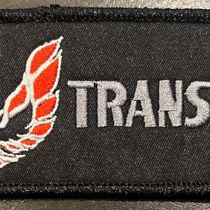 May include: Black rectangular patch with a stitched border. The patch features the words "TRANS AM" in gray, embroidered lettering. A red and white emblem is on the left side of the patch.