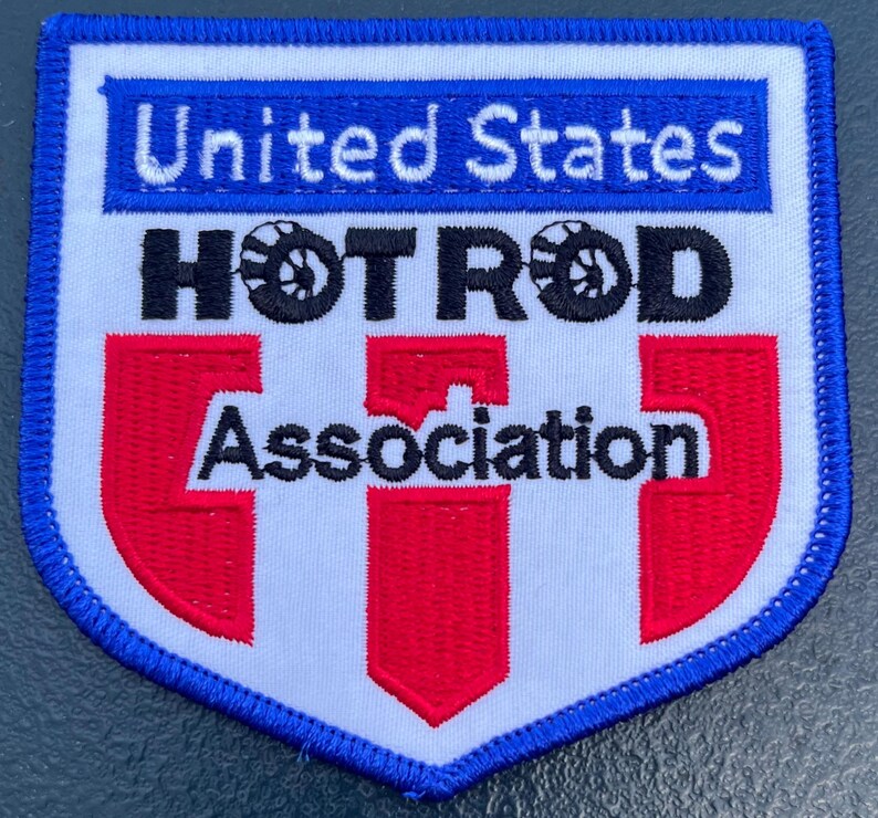 May include: A shield-shaped embroidered patch with a white background and a blue border. It features the text United States in white on a blue bar, the black text HOT ROD with tire icons, and the black text Association over three red vertical stripes.