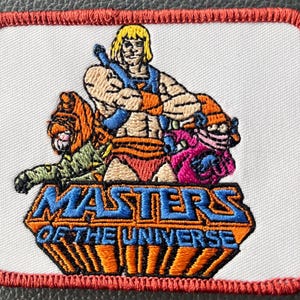 May include: Embroidered patch featuring He-Man, Battle Cat, and other characters from Masters of the Universe. The patch has a white background with a red border and the words "Masters of the Universe" in a blue and orange design.