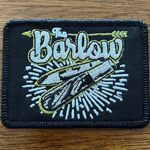 May include: Black embroidered patch with white and green stitching. The patch features the text "The Barlow" with an arrow and wheat stalks. The patch also features two stylized knives with a sunburst design.