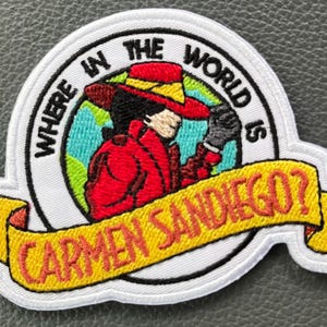 May include: Embroidered patch featuring a cartoon image of Carmen Sandiego in a red coat and hat. The patch has a white border with the text "WHERE IN THE WORLD IS CARMEN SANDIEGO?" on a yellow banner.