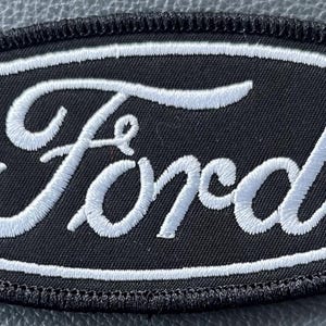 May include: Black oval patch with the white embroidered word "Ford". The patch has a white border and a black stitched edge. The patch is likely for a jacket or bag.