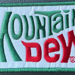 May include: Embroidered patch featuring the words "MOUNTAIN DEW" in green and red. The patch has a white background with a green border. The Mountain Dew logo is a popular beverage brand.
