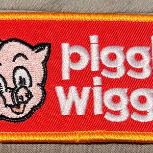 May include: Embroidered patch featuring a smiling pig wearing a chef's hat. The patch is red with white text that reads "piggly wiggly" and a yellow border. The pig is pink with black details.
