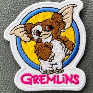 May include: Embroidered patch featuring a Gremlin character with brown fur, white chest, and large ears. The character is set against a yellow circle with a blue border, and the word "GREMLINS" is in pink letters.