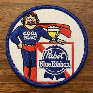 May include: A round, blue-bordered patch with a white background. A cartoon superhero figure wearing a blue suit with a red cape and the words "Cool Blue" on his chest is holding a glass of beer. The superhero is standing in front of a red and white box with the Pabst Blue Ribbon logo.