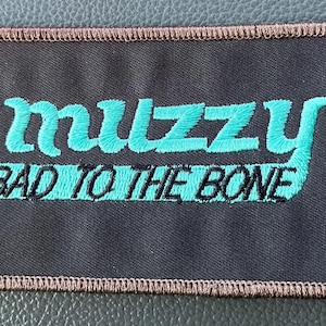 May include: Rectangular black patch with the word "muzzy" in turquoise script above the words "BAD TO THE BONE" in turquoise and black block letters. The patch has a brown stitched border.
