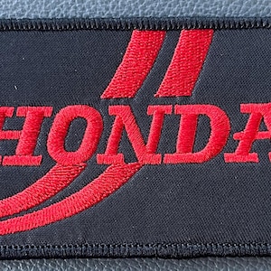 May include: Black rectangular patch with the red embroidered word "HONDA" and two red curved stripes. The patch has a black border with a stitched edge. The patch is likely for a jacket or bag.