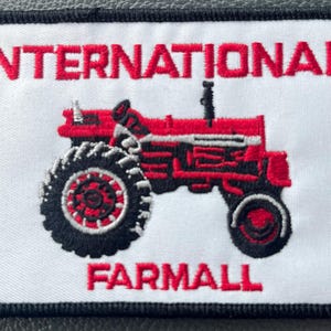 May include: Embroidered patch featuring a red International Farmall tractor on a white background. The words "INTERNATIONAL" and "FARMALL" are in red block letters above and below the tractor image. The patch has a black border.