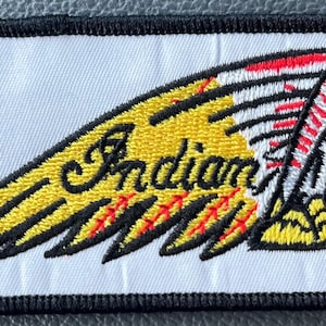 May include: Embroidered patch featuring the word "Indian" in a yellow wing design. The patch includes a red and white Native American profile. The patch has a black border and is on a white background.