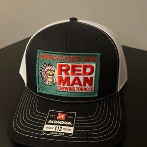 May include: Black and white trucker hat with a patch that reads "America's Best Chew Red Man Chewing Tobacco." The patch features a Native American chief graphic. The hat is adjustable and has a Richardson tag.