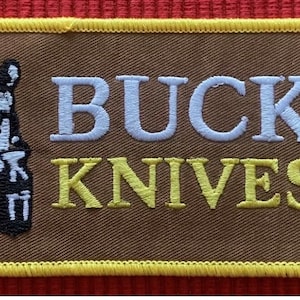 May include: A brown embroidered patch with a yellow border. The patch features a silhouette of a person holding a knife and the text "BUCK KNIVES" in white and yellow lettering.